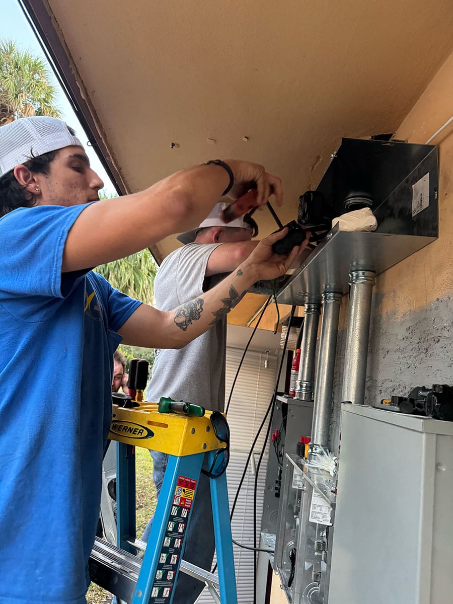 TNS Electric electricians stand on ladders installing or repairing electrical equipment on the exterior wall of a building.