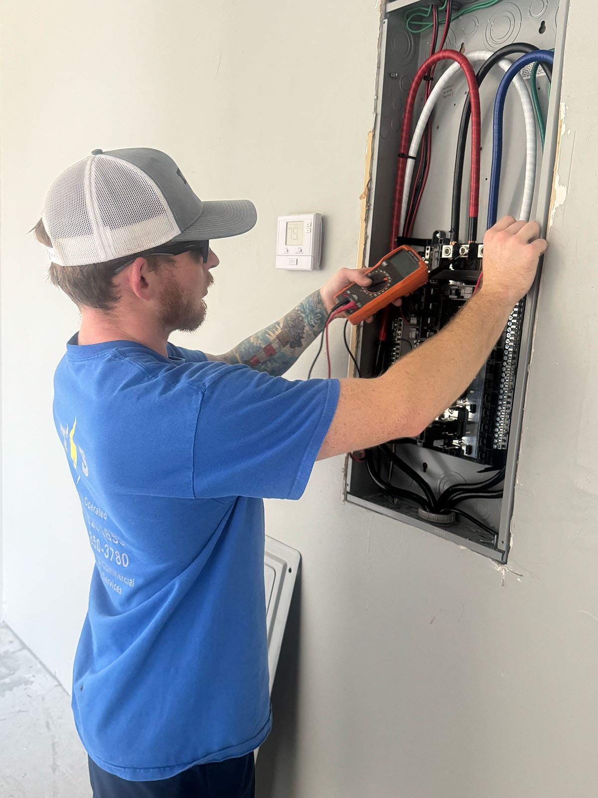 TNS Electric An electrician in a blue shirt and cap uses a multimeter to test wires in an open electrical panel mounted on a wall.